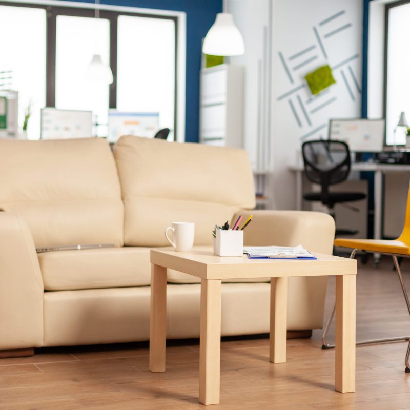 Modern business relax zone interior with confortable couch and orange chair. Rest area in company center with small sofa, shot of empty room with modern furniture, white shelves and blue wall.